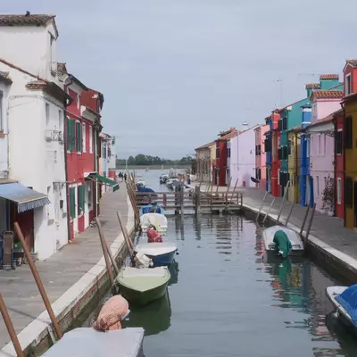 Canal in Burano lined with brightly coloured houses, small boats moored along both banks, the lagoon visible in the distance