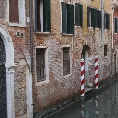 Venetian canal lined with aged brick buildings, an ornate wrought iron gate on the left and red-and-white striped mooring poles reflected in the green water