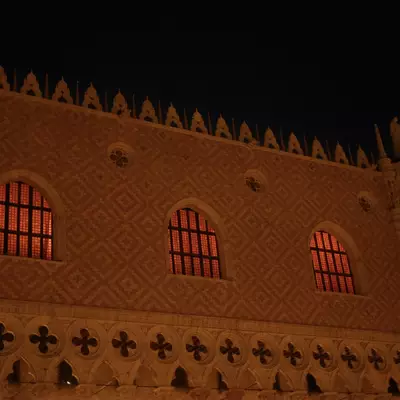 Upper facade of the Doge's Palace at night, warm amber light illuminating the Gothic arched windows and diamond-pattern stonework with decorative battlements along the roofline