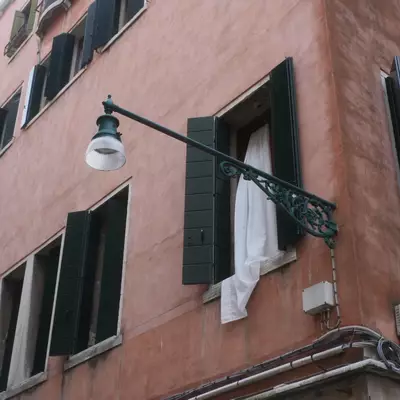 Low-angle view of a Venetian building facade with dark green shutters and a decorative iron street lamp bracket, a white curtain hanging from an open window