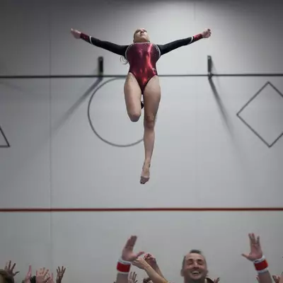 Female gymnast in dark red leotard thrown high into the air by teammates, arms spread wide, caught in mid-flight