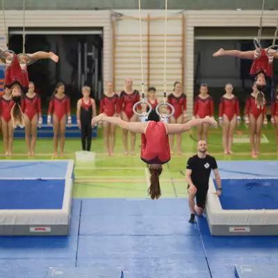 Three gymnasts in red leotards simultaneously performing identical inverted spread-eagle positions on rings with arms wide open, a coach watching from below, and a long line of teammates observing before a large crowd in the stands.