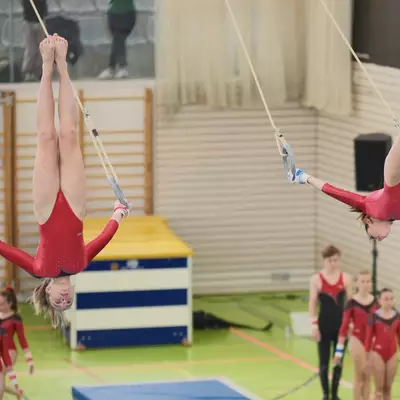 Two female gymnasts in red leotards simultaneously performing fully inverted positions on gymnastic rings, legs raised high in the air, with other gymnasts and spectators visible in the background.