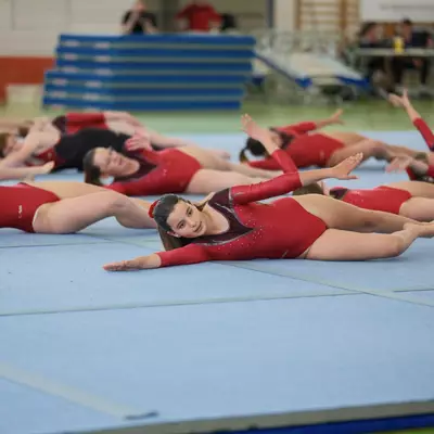 A group of gymnasts all lying flat on the floor mat in a synchronized side-lying position with arms extended, one gymnast in the center looking up toward the camera with a calm expression.