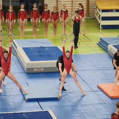 Three female gymnasts in red leotards simultaneously performing ring exercises, each supported by a coach in black, with a long line of teammates watching from the background.