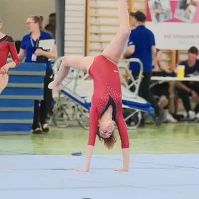 A young female gymnast in a red leotard performing a one-handed cartwheel on the floor mat, inverted with one leg extended high, while judges and officials sit in the background.