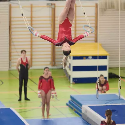A female gymnast in a red leotard hanging upside down from gymnastic rings, gripping the rings with both hands above her, with a coach and teammates visible below and spectators through a glass partition above.