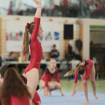 A female gymnast in a red leotard with a red flower hair accessory raises one arm with a smile during a floor exercise, while other gymnasts crouch in various poses behind her and spectators fill the stands above.