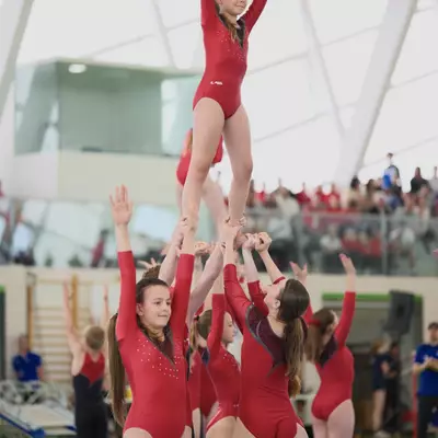 A group of gymnasts forming a human tower: one gymnast stands elevated on the hands and shoulders of several teammates with arms raised in triumph, all in red leotards, with a packed audience visible in the background.