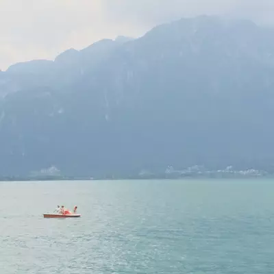 A small pedal boat carrying three people drifts on the misty waters of Lake Geneva, dwarfed by the towering silhouette of the Grammont massif and a lakeside town in the haze beyond.