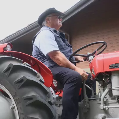 An older man in a wide-brimmed hat and sunglasses sits at the wheel of a vintage red Massey-Ferguson tractor, seen in profile in front of a chalet.