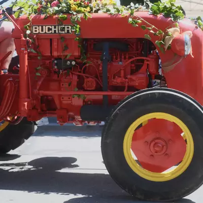 A vintage red Bucher tractor decorated with greenery, flowers and burlap ribbons, parked at a street parade with onlookers gathered alongside it.
