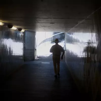 Silhouette of a man in a wide-brimmed hat walking away through a dim concrete underpass, heading toward bright daylight at the far end.