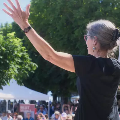 Seen from behind, a woman with grey hair in a bun raises both arms toward a sunlit crowd gathered under white tents along the lakeside.