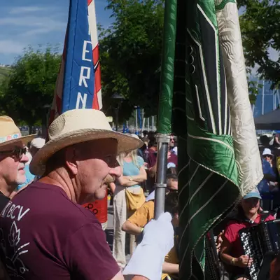 Two men in straw hats carry ceremonial flags through a festive lakeside crowd, with vineyard-covered hills visible across the water in the background.