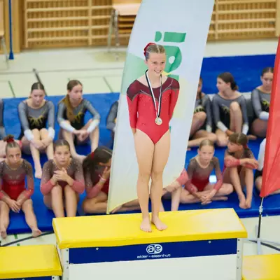 Young gymnast in a red leotard smiling on a yellow award podium, wearing a silver medal, sponsor banners and other gymnasts seated behind.
