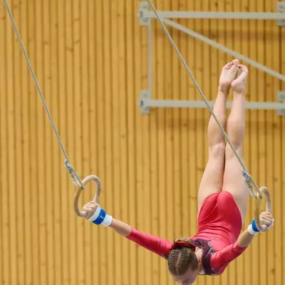 Gymnast in a red leotard performing a tucked inverted hang on gymnastics rings, body curled in a tight ball, blue crash mats below.