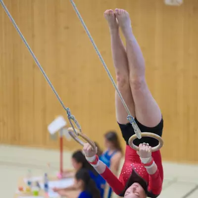 Gymnast in a red leotard performing an inverted piked position on gymnastics rings, legs pointing straight up, wooden gymnasium wall behind.