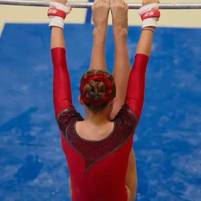 Gymnast in a red leotard seen from behind, hanging from the uneven bar with legs raised and feet near the bar, red flower in her hair bun, blue crash mat below.