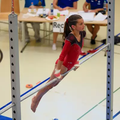 Young gymnast in a red leotard gripping the uneven bar with one arm, legs dangling, looking sideways, judges at a table visible in the background.