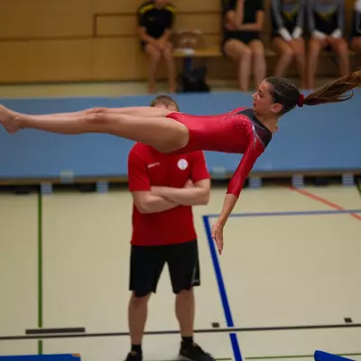 Gymnast in a red leotard performing a back layout in a straddle position, coach in a red shirt with arms crossed watching from below, other gymnasts seated on bleachers behind.