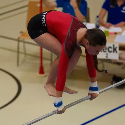 Gymnast in a red leotard crouching on the low uneven bar, gripping it with both hands and feet, judges seated at a table in the background.