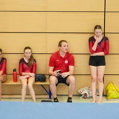 Two gymnasts in red leotards and their coach in a red polo shirt sitting on a bench watching the competition, another gymnast standing to the right.