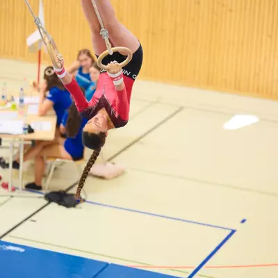 Gymnast in a red leotard hanging upside down on gymnastics rings with legs split wide, long braided hair hanging toward the ground, judges at tables visible in the background.