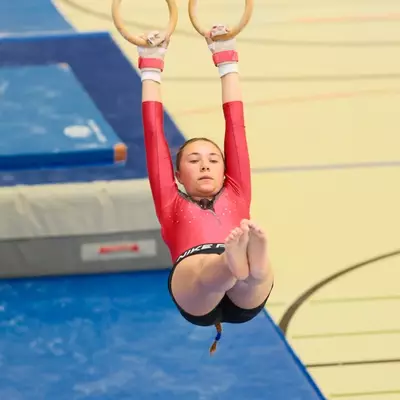 Gymnast in a red leotard hanging from gymnastics rings in a tucked pike position with knees bent and feet forward, blue crash mats below.