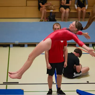 Gymnast in a red leotard doing a back walkover over a standing coach in a red shirt, her hair fanning out in mid-motion, other athletes visible in the background.