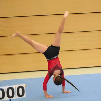 Gymnast in a red leotard performing a one-arm cartwheel on the floor mat, one leg raised high, a scoreboard showing 