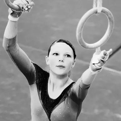 Gymnast in a red leotard hanging from gymnastics rings with knees raised to face height, looking upward, blue crash mats below.