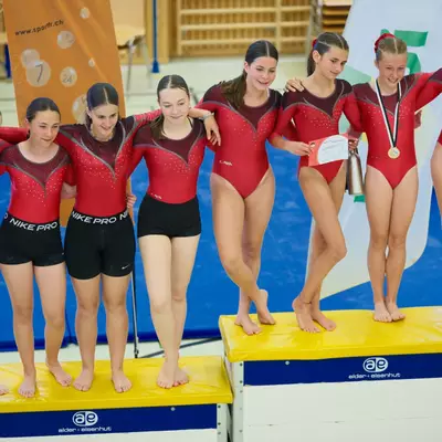 Group of seven Gym Ursy gymnasts in red leotards posing on yellow award podiums, one holding a trophy and another wearing a gold medal, sponsor banners visible in the background.
