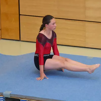 Gymnast in a red leotard seated on a blue floor mat with one leg extended, looking to the side, at the start or end of a floor routine.