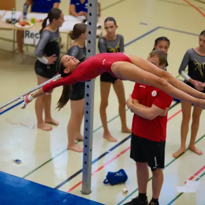 Gymnast in a red leotard performing a release move on uneven bars, arching backward over the bar with arms extended, coach watching with crossed arms in the background.