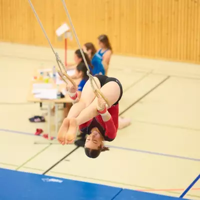 Gymnast in a red leotard performing an inverted tuck on gymnastics rings, body in a tight ball with knees bent close to the chest, wooden gymnasium wall behind.