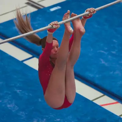 Gymnast in a red leotard swinging on the bar in a tight pike with feet raised toward her hands, hair sweeping outward, blue crash mat below.
