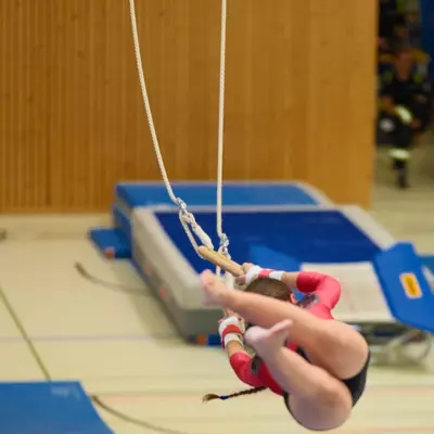 Gymnast in a red leotard performing a tight tuck on gymnastics rings, body curled close to the rings, vault apparatus and crash mats visible in the background.