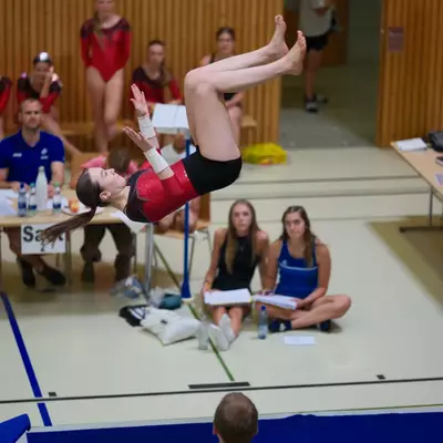 Gymnast in a red leotard mid-back salto above the floor mat with legs fully extended, judges seated at tables and other gymnasts watching in the background.