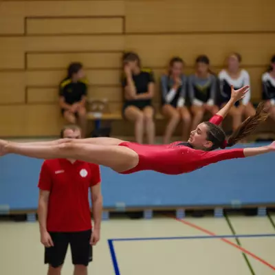 Gymnast in a red leotard performing a horizontal layout with arms spread wide in mid-air, coach watching in the background with other gymnasts on bleachers.