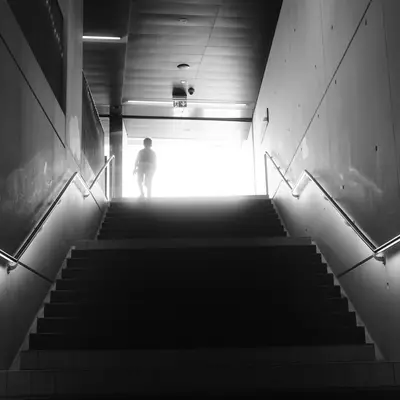 A person's silhouette at the top of a staircase, backlit by an intense white light flooding through an exit, in a underground passage with illuminated metal handrails on both sides.