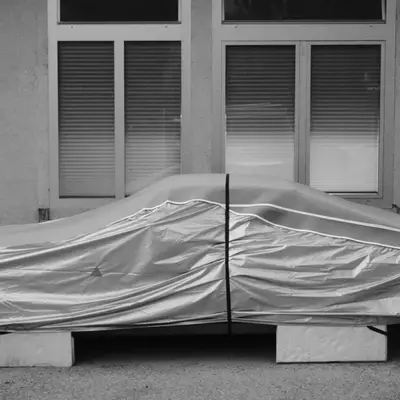 A car covered with a silver protective tarp, secured with a vertical strap, resting on concrete blocks in front of a building with shuttered windows