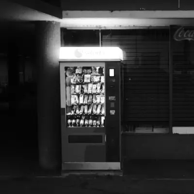 A Selecta vending machine glowing in the dark, next to a shuttered shop with a Coca-Cola sign and graffiti on a concrete pillar, photographed at night