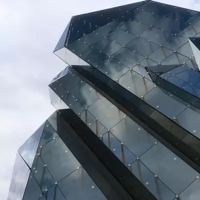 Low-angle view of a faceted, crystal-like building clad in dark reflective glass panels, its sharp angles set against a pale cloudy sky.