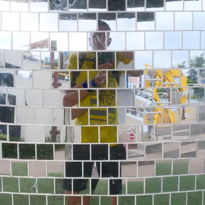 Self-portrait reflected in a curved wall of small square mirror tiles, showing a person in a yellow t-shirt and dark shorts holding up a camera in a theme park setting.