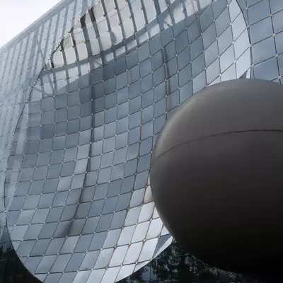 A large building covered in shimmering silver metallic scales, with a huge matte dark grey sphere rising in the foreground under an overcast sky.