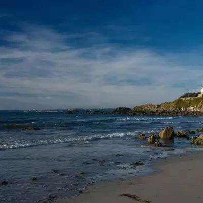 A sandy beach with dark rocks scattered along the shoreline and gentle waves rolling in, backed by a grassy hillside topped with a white castle or fortress structure under a partly cloudy blue sky.