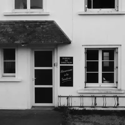 A white-walled cottage with a dark slate roof features a black door with a small porch, flanked by multi-pane windows. A bicycle rack sits against the right side of the building. A chalkboard sign hangs beside the entrance door.
