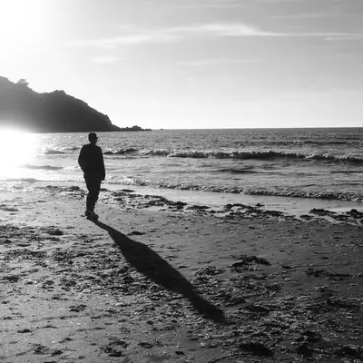 A person stands on a beach gazing at the ocean waves during golden hour. The black and white photograph captures a long shadow cast across the sandy beach, with forested cliffs visible in the background.