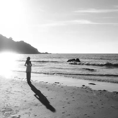 A person stands alone on a sandy beach at sunset or sunrise, their long shadow stretching across the wet sand toward the camera, with gentle waves and a rocky coastline visible in the background.
